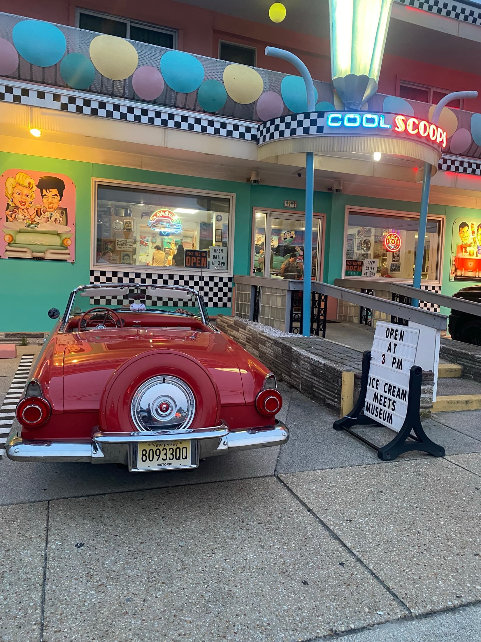 The Cool Scoops storefront at dusk, neon sign lit, with a red 1950s Thunderbird parked out front